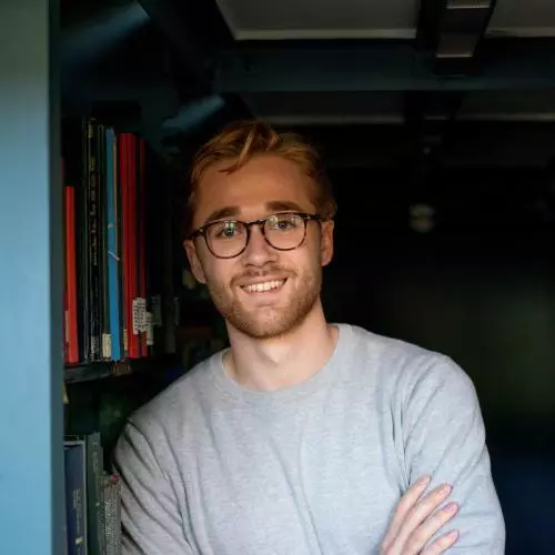 Robert Hill has short fair hair and is wearing round tortoiseshell glasses and a grey jumper. He is leaning against a bookshelf.