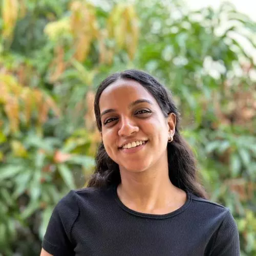 Tejashwari has long dark hair. She is wearing a black short sleeved t-shirt. She is standing in front of some foliage, smiling. 