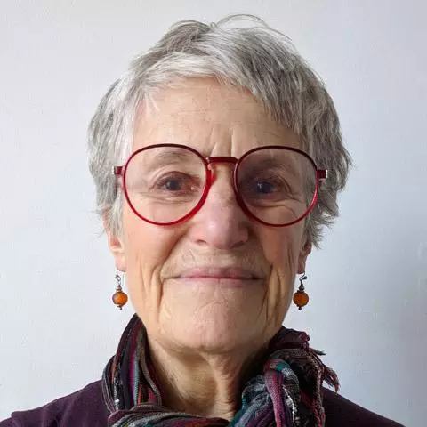 Woman with short grey hair, smiling and wearing red rimmed glasses and scarf around her neck.