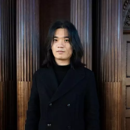 Xinghao li is wearing a black coat, with shoulder length hair looking at the directly at the camera.