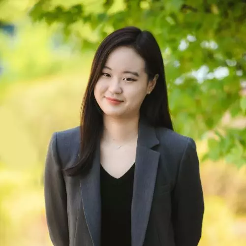 Ye Eun Ha has long dark hair and is wearing a black top and grey jacket. She's standing in front of a tree