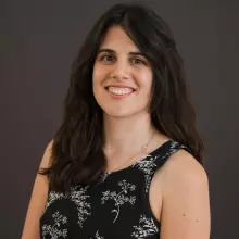 Woman with long brown hair, smiling at the camera and wearing a black dress with white pattern.