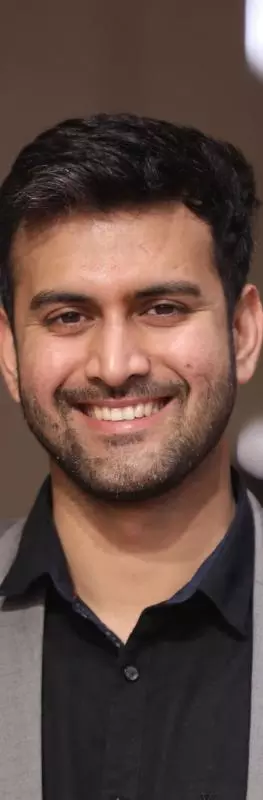 Sayyed has short black hair. He is wearing a black shirt and grey blazer