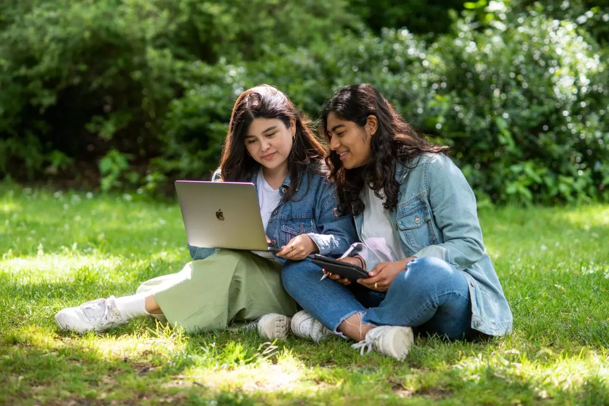 Two students sitting close together on some grass. The student on the left is holding a laptop on her knee. The other student is leaning to take a look at the screen.